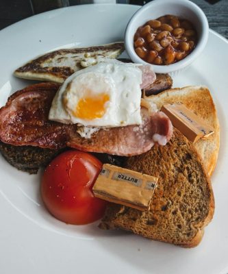 From above of toasted bread with fried bacon and egg served on white table with tomato and beans on wooden table